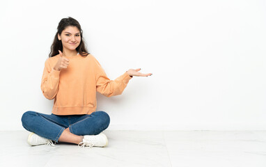 Wall Mural - Teenager Russian girl sitting on the floor holding copyspace imaginary on the palm to insert an ad and with thumbs up