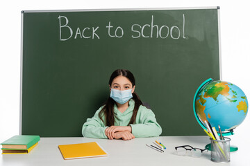 Wall Mural - Schoolkid in medical mask sitting near books and chalkboard with back to school lettering isolated on white.