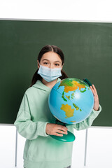 Wall Mural - Pupil in medical mask holding globe near chalkboard isolated on white.