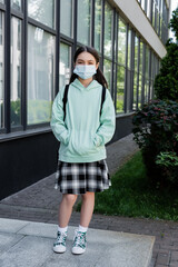 Wall Mural - Schoolchild in medical mask standing near building on urban street.