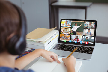 Sticker - Caucasian boy writing in book while learning through online class over video on laptop at home