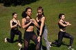 Fit sporty caucasian female fitness trainer helping african woman learning lunge on group outdoor class. Instructor and multiethnic ladies do workout training exercise on green grass in park outside.