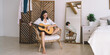 © BullRun - Brunette attractive female smiling and learning to play guitar while sitting in cozy home interior. Young happy woman enjoying her hobby with musical instrument performance relaxing on wicker chair