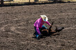 A rodeo cowboy in a white hat and pink shirt has a calf on the ground beginning to tie up its legs in a calf roping competition. The arena is dirt.