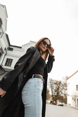 Wall Mural - Stylish beautiful young woman in a fashionable black leather long coat with high-waisted jeans and a tank top is fixing cool sunglasses in the city. Gorgeous female urban style dress