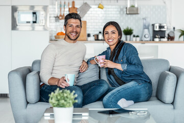 Lovely couple holding coffee cups while looking at camera sitting on couch at home.