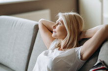 Satisfied Mature Caucasian Woman Is Relaxing On The Sofa At Living Room At Home. Middle Aged Blonde Woman Enjoying Weekend Or Leisure, Sits On Sofa, Looks Away And Dreams About Vacation