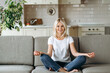 © Kateryna - Smiling caucasian middle aged female sits on sofa at living room and  practise yoga, meditation in lotus position looks at camera and smiling, relieve stress