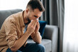 © Kateryna - Sick caucasian guy sits on couch feel unhealthy, he suffering from rhinitis snuffles, having respiratory infection, get flue,  sitting at home at quarantine
