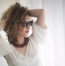 Slim Young Fashion Model With Curly Hair Wearing White Blouse In A Frame Of Window. Picture Of Lovely Sexy Fashionable Woman With Sunglasses And Necklace