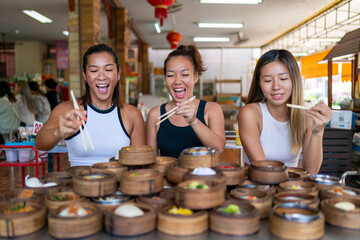 Wall Mural - Group of Asian woman tourist eating Chinese food steamed dumpling in bamboo steamer with chopsticks in Chinese restaurant. Happy female friends enjoy eating and travel together on summer vacation.