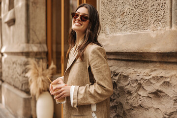 Fashionable young caucasian brunette girl with happy smile posing outdoors. Model wears sunglasses, beige jacket, holds glass. Concept of enjoying moment