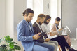 © Studio Romantic - Group of serious people waiting for job interview or business appointment in office. Four men and women sitting in line, holding paper resumes, going over reports, using modern tablets and laptops