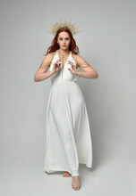 Full Length Portrait Of Beautiful Red Head Woman Wearing Long Flowing Fantasy Toga Gown With Golden Halo Crown Jewellery, Standing Pose   Isolated On A White Studio Background.

