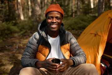 Happy african american man in spring coat and hat sitting next to orange tent in campsite in the middle of wild forest, enjoying fresh air and tranquil morning, holding thermos mug and smiling