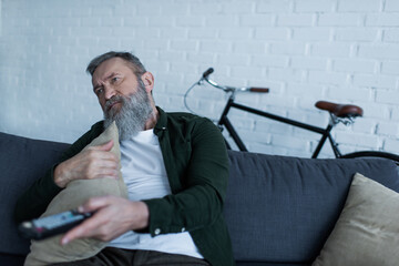 Wall Mural - depressed senior man holding remote controller and pillow while watching tv in living room.