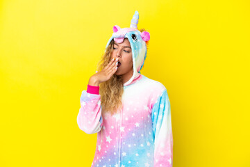 Wall Mural - Girl with curly hair wearing a unicorn pajama isolated on yellow background yawning and covering wide open mouth with hand