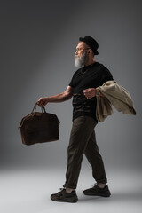 Poster - full length of stylish senior man in derby hat holding leather bag and beige jacket on grey.