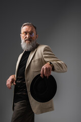 Poster - elegant senior man in beige jacket and eyeglasses holding derby hat on grey.