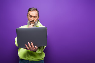 Wall Mural - pensive senior man with beard and grey hair using laptop on purple background.