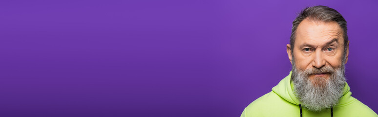 Wall Mural - skeptical senior man moving eyebrow while looking at camera on purple background, banner.