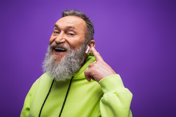 Wall Mural - cheerful senior man in green hoodie listening music and pointing with finger at wireless earphone on purple.