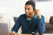 © kucherav - African american young man customer support call center operator or receptionist sitting at the workplace in a modern office consulting a client, uses a headset, smiles friendly