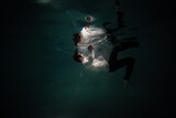 Fototapeta Sport - Sink. A young guy in a white shirt falls into the water, a photo from under the water. The concept of falling down, diving to the depth, contrasting dark photo