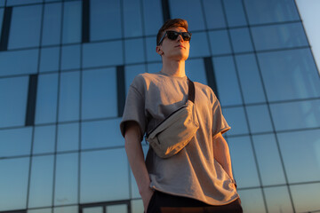 Wall Mural - Portrait of handsome tourist hipster man in sunglasses with bag handy, standing guy near the building in downtown. Calm relaxing moment, summer vacation concept