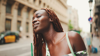 Wall Mural - Gorgeous woman with African braids wearing top stands on the sidewalk next to the road waiting for taxi