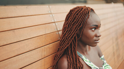 Wall Mural - Profile gorgeous woman with African braids wearing in top and trousers is resting sitting on bench