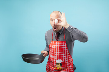 A chef man in red kitchen apron holding frying pan and juggling raw egg isolated on blue background.