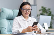 © Liubomir - Young beautiful Asian business woman works in modern office, uses phone, smiles, reads good news, chats in app with friends