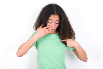 Wall Mural - Shocked Teenager girl with afro hairstyle wearing green T-shirt over white wall look surprisedly down, indicates at blank  with fore finger, Scared model