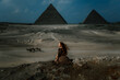 Young redhead tourist girl in brown dress sitting on a stone cliff in Egypt, Cairo - Giza. Pyramids on backround and dramatic light. Copy space