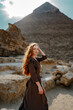 Young redhead tourist girl in brawn dress with a leather backpack standing on the sand in Egypt, Cairo - Giza. Pyramid of Khafre on backround. Sunset