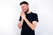 © Jihan - Overjoyed young caucasian bearded man wearing black t-shirt standing over white wall laughs joyfully and keeps palms pressed together hears something funny