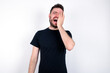 © Jihan - young caucasian bearded man wearing black t-shirt standing over white wall makes face palm and smiles broadly, giggles positively hears funny joke poses