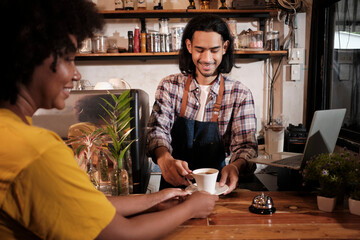 Wall Mural - In front close-up, A young long hair Thai male cafe barista works by serving a cup of coffee to African American female customer at counter bar of coffee shop, happy service job, and SME entrepreneur.