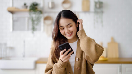 Wall Mural - Happy young asian woman relaxing at home. Asia female standing at counter kitchen and using mobile smartphone