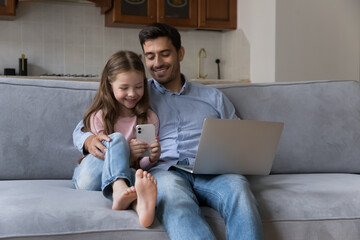 Cute preschool daughter holding smartphone sit on sofa with young father use laptop, family using diverse devices spend leisure on internet. Young generation and gadgets overuse, apps usage concept