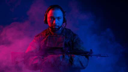 a beefy man in headphones and military clothes holds a machine gun with two hands to his chest. colored smoke around. blue background, multicolored light. portrait