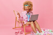 Joyful curly female freelance worker works online on laptop computer pretends being on resort during summer holidays poses on deck chair indicates left into distance notices something funny.