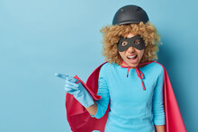 Displeased Irritated Woman With Curly Hair Dressed Like Superhero Indicates Left On Blank Space Shows Something Unpleasant Isolated Over Blue Background Invites You To Costume Party Or Masquerade