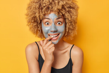 Wall Mural - Positive surprised European woman uses new beauty product applies clay mask on face to cleanse smooth and brighten skin bites finger wears black t shirt isolated over vivid yellow background