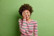 Happy curly haired woman makes face palm smiles gladfully expresses positive emotions keeps eyes closed dressed in casual striped jumper isolated over vivid green background hears something funny