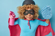© WHstudio Leushin N - Powerful serious woman superhero holds spray detergent and mop dressed in costume maintains cleaning service focused attentively at camera isolated over blue background. Housekeeping concept