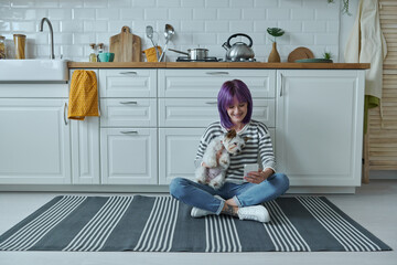 Wall Mural - Young woman carrying little dog and using smart phone while sitting on the floor at the kitchen