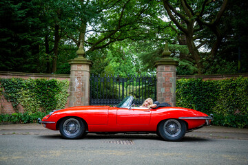 Lady with long blond hair resting in a red E-Type Jaguar with gates and park in the background