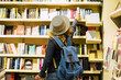 © simona - Back view of woman with hat and backpack choosing a book to buy inside a library or newspaper store. People prepare to travel. Leisure and shopping female people activity. Traveler lifestyle